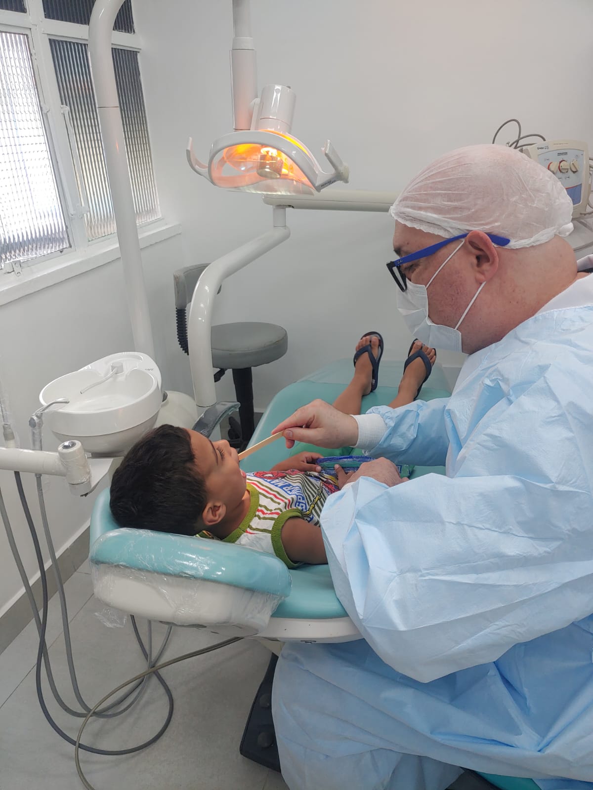 A foto mostra um dentista, visto de lado, examinando a boca de um menino de cerca de seis anos